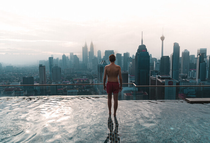 A man stands in the shallow area of an infinity pool, gazing out at the hazy, modern urban scenery, creating a tranquil and contemplative scene.